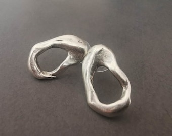 May include: A pair of silver-toned stud earrings featuring an irregular, organic, open-loop shape with a smooth, reflective metallic surface. Each earring has a rounded, asymmetrical form with a hollow center and a clear plastic backing.