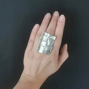 May include: A silver ring with a textured, rectangular design. The ring is worn on a person's finger.