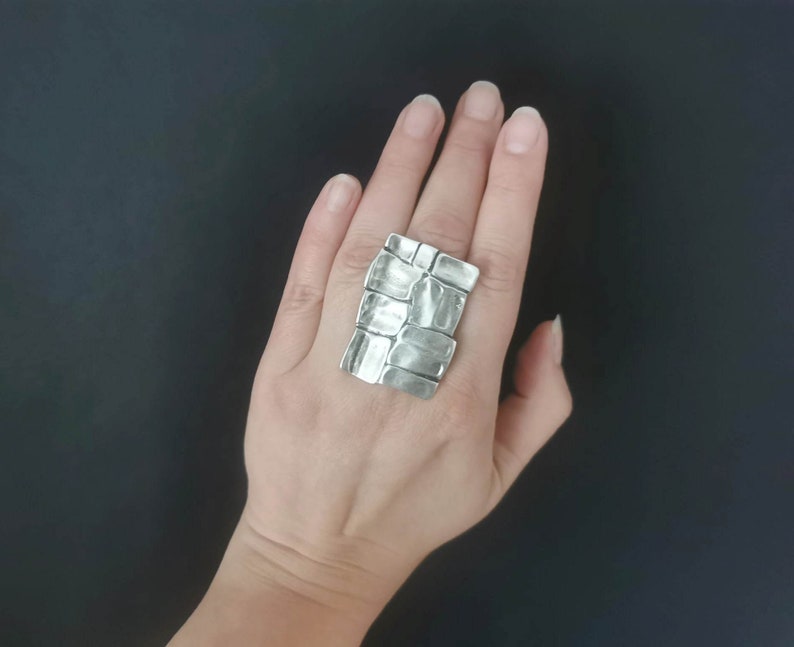 May include: A silver ring with a textured, rectangular design. The ring is worn on a person's finger.