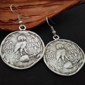 May include: Silver tone owl earrings with a Greek inscription. The earrings are round and feature a detailed owl design.