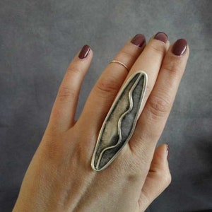 May include: A silver ring with a long, narrow, abstract design. The ring is worn on the index finger of a hand with dark red nail polish.