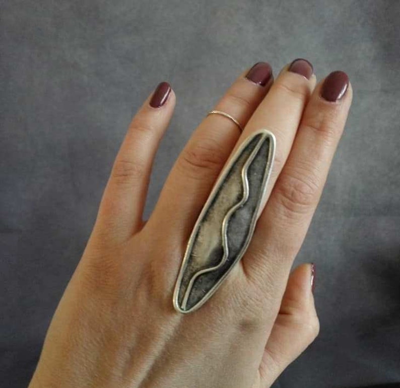May include: A silver ring with a long, narrow, abstract design. The ring is worn on the index finger of a hand with dark red nail polish.
