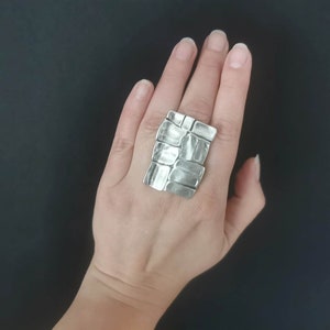 May include: A silver ring with a textured, rectangular design. The ring is worn on a person's finger.