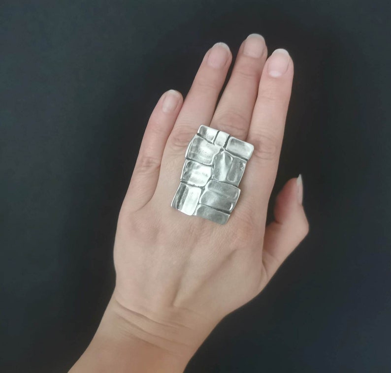 May include: A silver ring with a textured, rectangular design. The ring is worn on a person's finger.