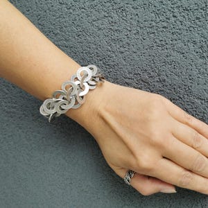 May include: A silver-toned bracelet with a linked circle design is worn on a wrist. A matching ring is on the finger. The jewelry is displayed against a gray background.