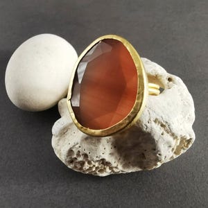 Peut inclure: Une bague dor&eacute;e orn&eacute;e d'une grande pierre pr&eacute;cieuse ovale, facett&eacute;e, brun-rouge. La bague est pos&eacute;e sur une pierre textur&eacute;e blanc cass&eacute;, avec une pierre ovale blanche et lisse en arri&egrave;re-plan. Le sertissage de la bague est martel&eacute;.