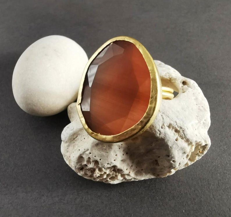 Peut inclure: Une bague dor&eacute;e orn&eacute;e d'une grande pierre pr&eacute;cieuse ovale, facett&eacute;e, brun-rouge. La bague est pos&eacute;e sur une pierre textur&eacute;e blanc cass&eacute;, avec une pierre ovale blanche et lisse en arri&egrave;re-plan. Le sertissage de la bague est martel&eacute;.