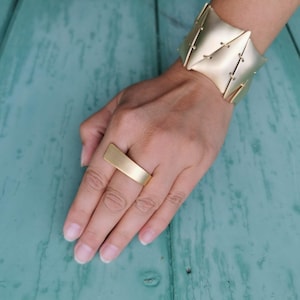 Op de afbeelding: Goudkleurige sieradenset met een brede manchetarmband en een rechthoekige ring. De manchet heeft een geometrisch ontwerp met gestikte accenten. De ring is een eenvoudige, platte band. De sieraden worden getoond op een turquoise houten oppervlak.