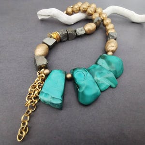 Puede incluir: Un collar artesanal con una combinación de cuentas doradas, cubos de pirita y grandes piedras irregulares de malaquita turquesa. Una cadena dorada cuelga del collar, añadiendo un toque de elegancia. El collar se muestra sobre una superficie gris oscuro.
