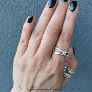 Może przedstawiać: Ręka z srebrnym pierścionkiem o falistym wzorze. Pierścionek jest na środkowym palcu. Na małym palcu jest również srebrny pierścionek. Paznokcie są pomalowane na czarno.