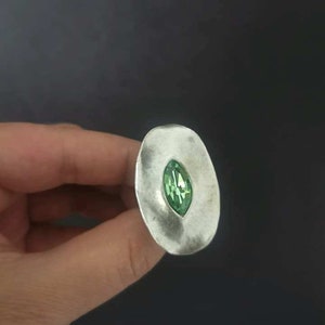 Puede incluir: Un anillo de plata con una piedra preciosa verde engastada en el centro. El anillo tiene forma ovalada y una superficie texturizada.