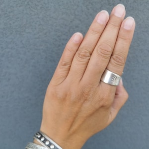 May include: A silver ring with a textured design on the band. The ring is worn on the ring finger of a person's hand.