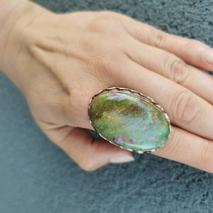 May include: An oval-shaped ring featuring a large, polished gemstone with a marbled surface of moss green, earthy brown, and small flecks of magenta. The stone is set within a simple, thin metallic band that follows the oval contour of the gem.