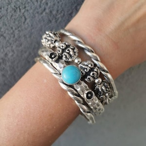 Peut inclure: Un empilement de quatre bracelets en métal argenté. L'un des bracelets présente une pierre turquoise ronde et lisse. Un autre a une texture torsadée, tandis que les autres affichent une finition martelée et de petites perles texturées.