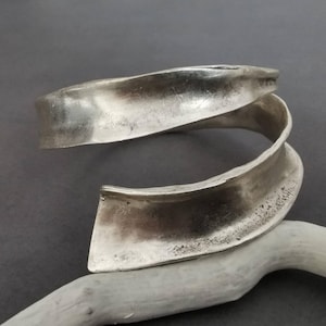 Può includere: Un bracciale a fascia color argento con un design aperto a spirale. La superficie metallica presenta una finitura opaca e testurizzata con variazioni visibili di tono e profondità, creando una forma organica curva che si avvolge su se stessa.