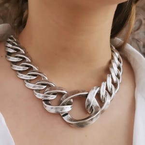 Puede incluir: Un collar de cadena plateada y gruesa con eslabones grandes y texturizados. El collar presenta un cierre central prominente y un acabado envejecido, creando una pieza audaz. El collar se lleva alrededor del cuello.