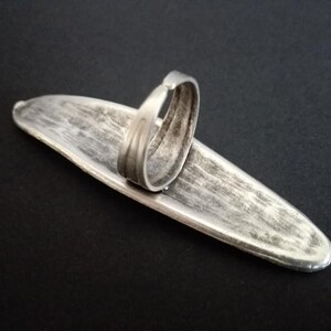 May include: A silver-toned ring with a wide, flat band. The band is oval-shaped and has a textured surface. The ring is designed to be worn on the finger.