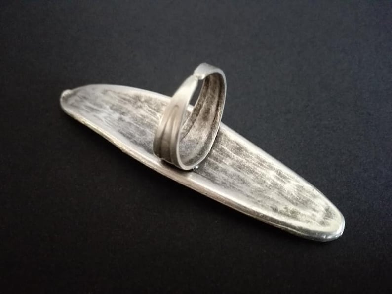 May include: A silver-toned ring with a wide, flat band. The band is oval-shaped and has a textured surface. The ring is designed to be worn on the finger.