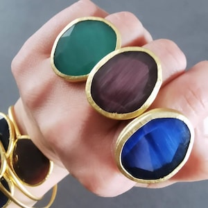 May include: Three gemstone rings with gold-colored bands are displayed. The rings feature large, oval-shaped stones in green, purple, and blue. A gold bracelet with a similar design is visible in the background. The rings are on a hand.
