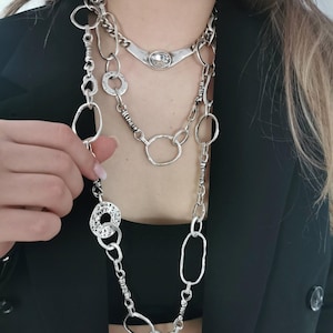 Puede incluir: Collar plateado de varias hebras con varios estilos de cadena y diseños de colgantes. El collar presenta un colgante grande de forma ovalada, eslabones circulares texturizados y una variedad de otras formas geométricas. El collar se usa sobre una blusa y un blazer negros.