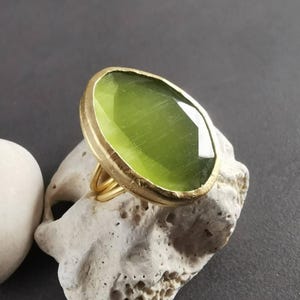 Peut inclure: Une bague dor&eacute;e orn&eacute;e d'une grande pierre pr&eacute;cieuse ovale vert clair. La pierre est sertie dans une lunette dor&eacute;e et repose sur une bague dor&eacute;e &agrave; plusieurs bandes. La bague est pr&eacute;sent&eacute;e sur une pierre blanche cass&eacute; textur&eacute;e sur fond gris fonc&eacute;.