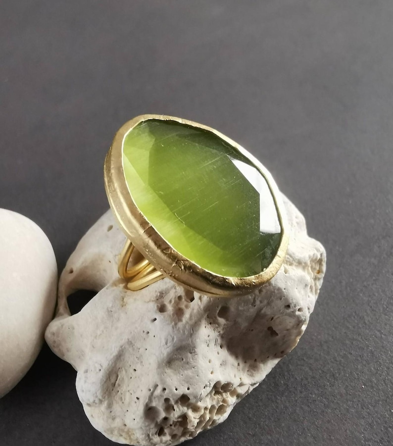 Peut inclure: Une bague dor&eacute;e orn&eacute;e d'une grande pierre pr&eacute;cieuse ovale vert clair. La pierre est sertie dans une lunette dor&eacute;e et repose sur une bague dor&eacute;e &agrave; plusieurs bandes. La bague est pr&eacute;sent&eacute;e sur une pierre blanche cass&eacute; textur&eacute;e sur fond gris fonc&eacute;.
