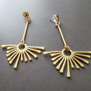 May include: A pair of gold-colored earrings. Each earring features a long, thin bar connected to a sunburst design with multiple rays. The earrings have a post-back closure and are displayed against a gray background.