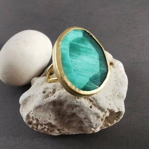 Peut inclure: Bague dor&eacute;e orn&eacute;e d'une grande pierre verte ovale et facett&eacute;e. La pierre est sertie dans un cadre dor&eacute; et repose sur une pierre textur&eacute;e blanc cass&eacute;. Le chaton de la bague est &eacute;galement dor&eacute;.