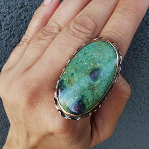Puede incluir: Un anillo ovalado grande con una piedra verde pulida que presenta inclusiones de color rojo oscuro. La piedra está engastada en una banda de metal festoneada con acabado metálico, mostrando un borde texturizado que enmarca la gema lisa.