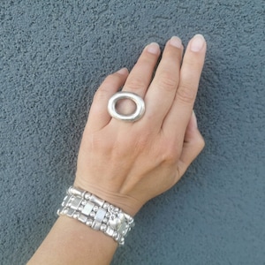Peut inclure: Une bague en argent avec une grande ouverture ovale. La bague est portée à l'index d'une main portant une pile de bracelets en argent.
