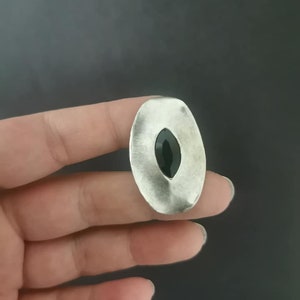 Pode incluir: Um pendente oval prateado com uma pedra preta no centro. O pendente tem uma superfície texturizada e uma pedra escura em forma oval no centro. A joia é segurada em uma mão contra um fundo escuro.