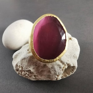 Peut inclure: Une bague imposante avec une pierre pr&eacute;cieuse magenta ovale sertie dans un cadre dor&eacute;. La pierre a une surface facett&eacute;e. La bague est pos&eacute;e sur une pierre blanche textur&eacute;e, avec une pierre blanche en arri&egrave;re-plan.