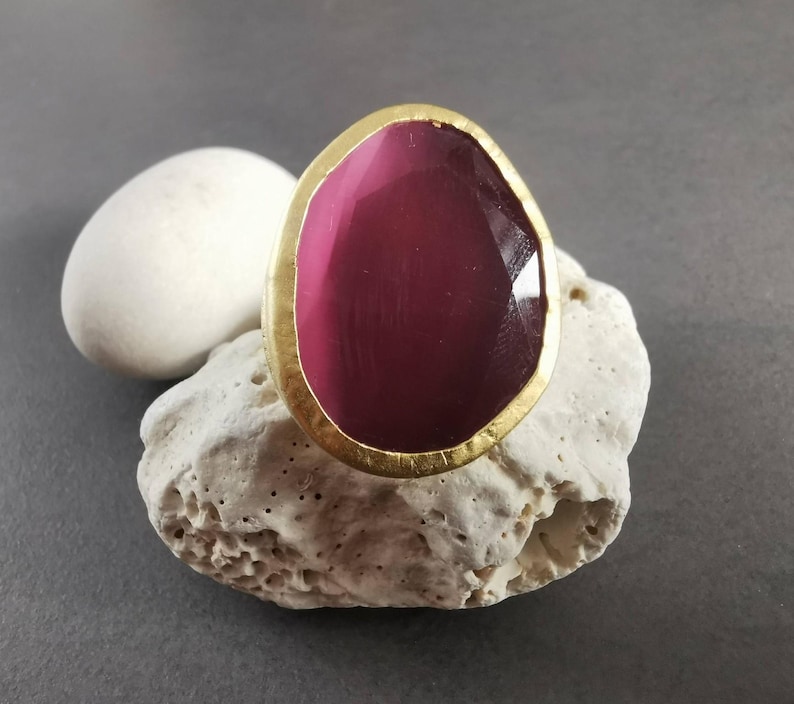 Peut inclure: Une bague imposante avec une pierre pr&eacute;cieuse magenta ovale sertie dans un cadre dor&eacute;. La pierre a une surface facett&eacute;e. La bague est pos&eacute;e sur une pierre blanche textur&eacute;e, avec une pierre blanche en arri&egrave;re-plan.
