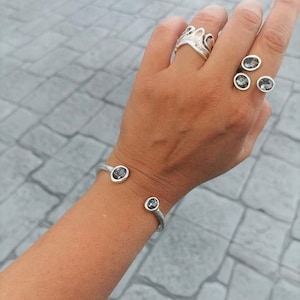Puede incluir: Una pulsera de plata con dos piedras preciosas negras. La pulsera está abierta y tiene un diseño sencillo. La pulsera se lleva en la muñeca de una persona.