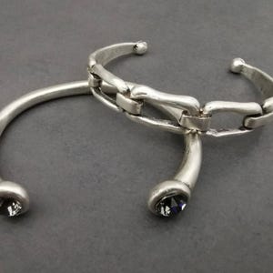 Może przedstawiać: Dwie bransoletki z metalu w kolorze srebrnym. Jedna to prosta bransoleta z ciemnym kamieniem na każdym końcu. Druga bransoletka ma wz&oacute;r ogniw łańcucha. Obie bransoletki są prezentowane na szarej powierzchni.