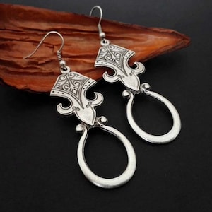 May include: A pair of silver earrings with an intricate design. Each earring has a decorative top section with swirling patterns and a teardrop-shaped bottom. The earrings are shown on a dark surface with a brown leaf in the background.