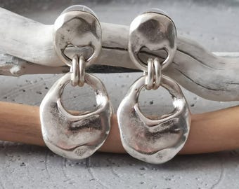 May include: A pair of silver-toned drop earrings featuring a circular top stud connected by two small rings to a larger, irregular, organic-shaped pendant. The metal surface has a hammered, textured finish with a reflective, polished appearance.