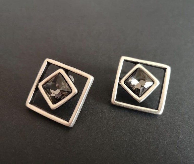 May include: A pair of silver earrings with a square frame and a dark gray crystal in the center.