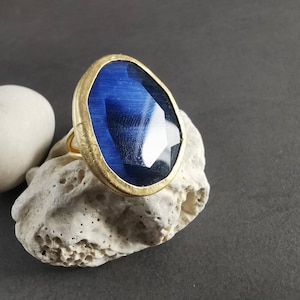 Peut inclure: Une bague dor&eacute;e orn&eacute;e d'une grande pierre pr&eacute;cieuse bleue ovale et facett&eacute;e. La bague est pos&eacute;e sur une pierre textur&eacute;e blanc cass&eacute;, avec une pierre blanche lisse en forme d'ovale &agrave; gauche. Le fond est gris fonc&eacute;.