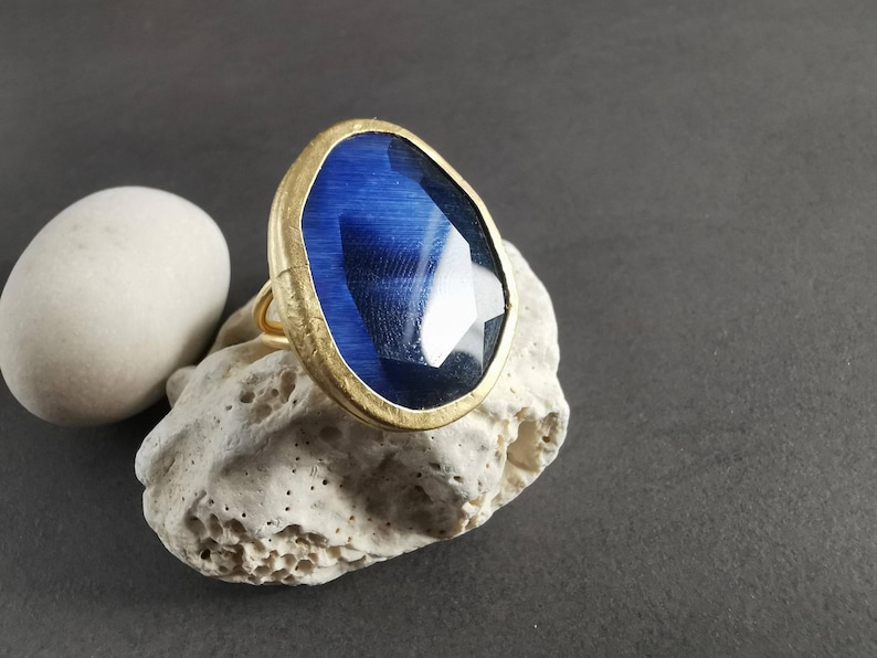 Peut inclure: Une bague dor&eacute;e orn&eacute;e d'une grande pierre pr&eacute;cieuse bleue ovale et facett&eacute;e. La bague est pos&eacute;e sur une pierre textur&eacute;e blanc cass&eacute;, avec une pierre blanche lisse en forme d'ovale &agrave; gauche. Le fond est gris fonc&eacute;.