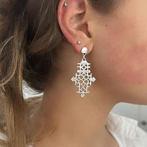 Può includere: Orecchini pendenti in argento con un design geometrico. Gli orecchini hanno la forma di una croce con dettagli intricati.
