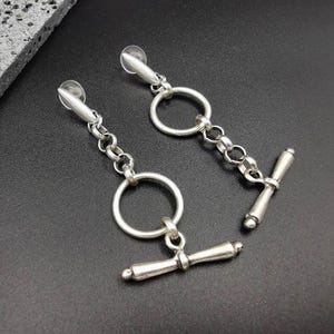 Può includere: Un paio di orecchini pendenti color argento. Ogni orecchino presenta un perno, una catena, un anello grande e una chiusura a barra a T. Gli orecchini sono esposti su una superficie nera.