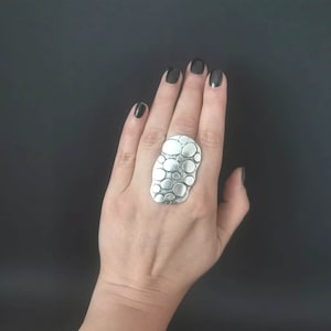 Peut inclure: Une bague en argent avec un motif texturé de nombreux petits cercles. La bague est portée sur une main avec un vernis à ongles foncé.