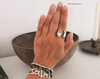 Puede incluir: Un anillo plateado con una gema clara de forma ovalada se muestra en una mano. El anillo tiene un diseño único con una banda abierta. Varias pulseras con cuentas plateadas y turquesas adornan la muñeca.
