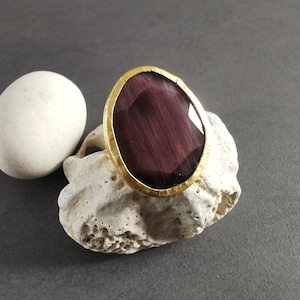 Peut inclure: Une bague dor&eacute;e avec une grosse pierre pr&eacute;cieuse ovale, violet fonc&eacute;. La pierre a un motif ray&eacute;. La bague est pr&eacute;sent&eacute;e sur un morceau de pierre blanche textur&eacute;e, avec une pierre blanche lisse &agrave; gauche.