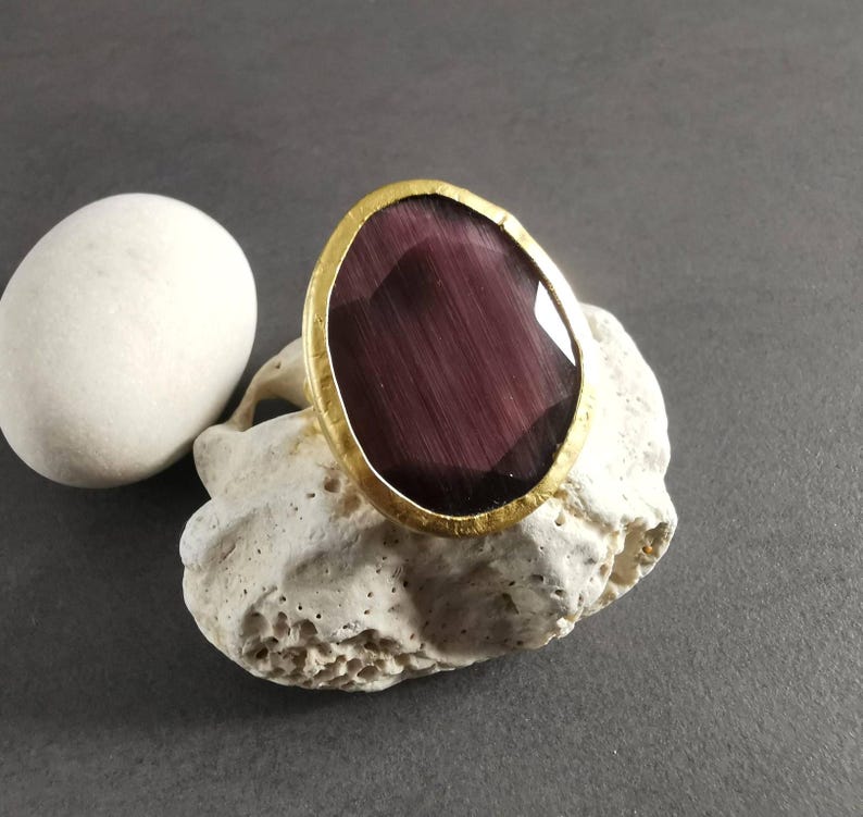 Peut inclure: Une bague dor&eacute;e avec une grosse pierre pr&eacute;cieuse ovale, violet fonc&eacute;. La pierre a un motif ray&eacute;. La bague est pr&eacute;sent&eacute;e sur un morceau de pierre blanche textur&eacute;e, avec une pierre blanche lisse &agrave; gauche.