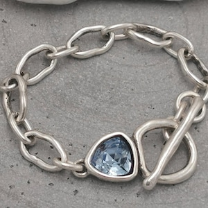 May include: A silver bracelet with a chain link design and a triangular, light blue gemstone. The bracelet features a toggle clasp and is displayed on a gray surface. The jewelry is a fashion accessory.