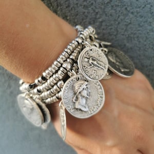 Può includere: Un bracciale color argento a più fili caratterizzato da numerose piccole perline metalliche e diversi ciondoli a forma di moneta. Ogni moneta presenta profili e iscrizioni in rilievo, creando un aspetto stratificato con una finitura metallica.
