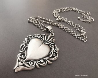May include: A silver necklace with a large, ornate heart pendant. The pendant is detailed with intricate scrollwork and has a smooth, heart-shaped center.