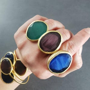 Peut inclure: Trois bagues orn&eacute;es de grandes pierres ovales vertes, violettes et bleues, serties dans des montures dor&eacute;es. Un bracelet dor&eacute; avec des d&eacute;tails de pierres assorties est &eacute;galement visible, sur fond gris.